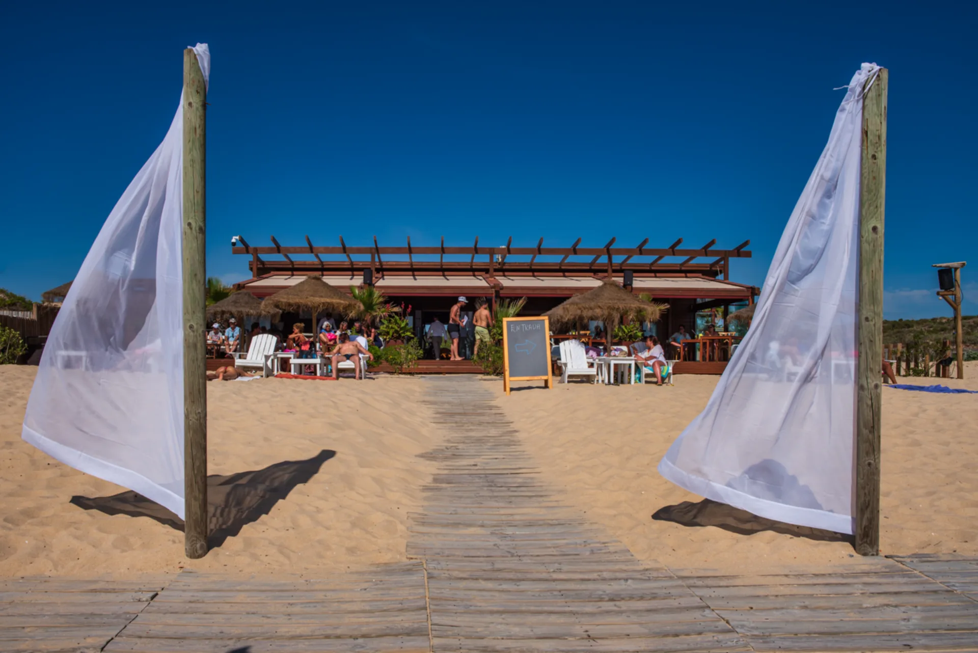 Boardwalk leading to a thatched beach bar with white fabric panels billowing in the breeze
