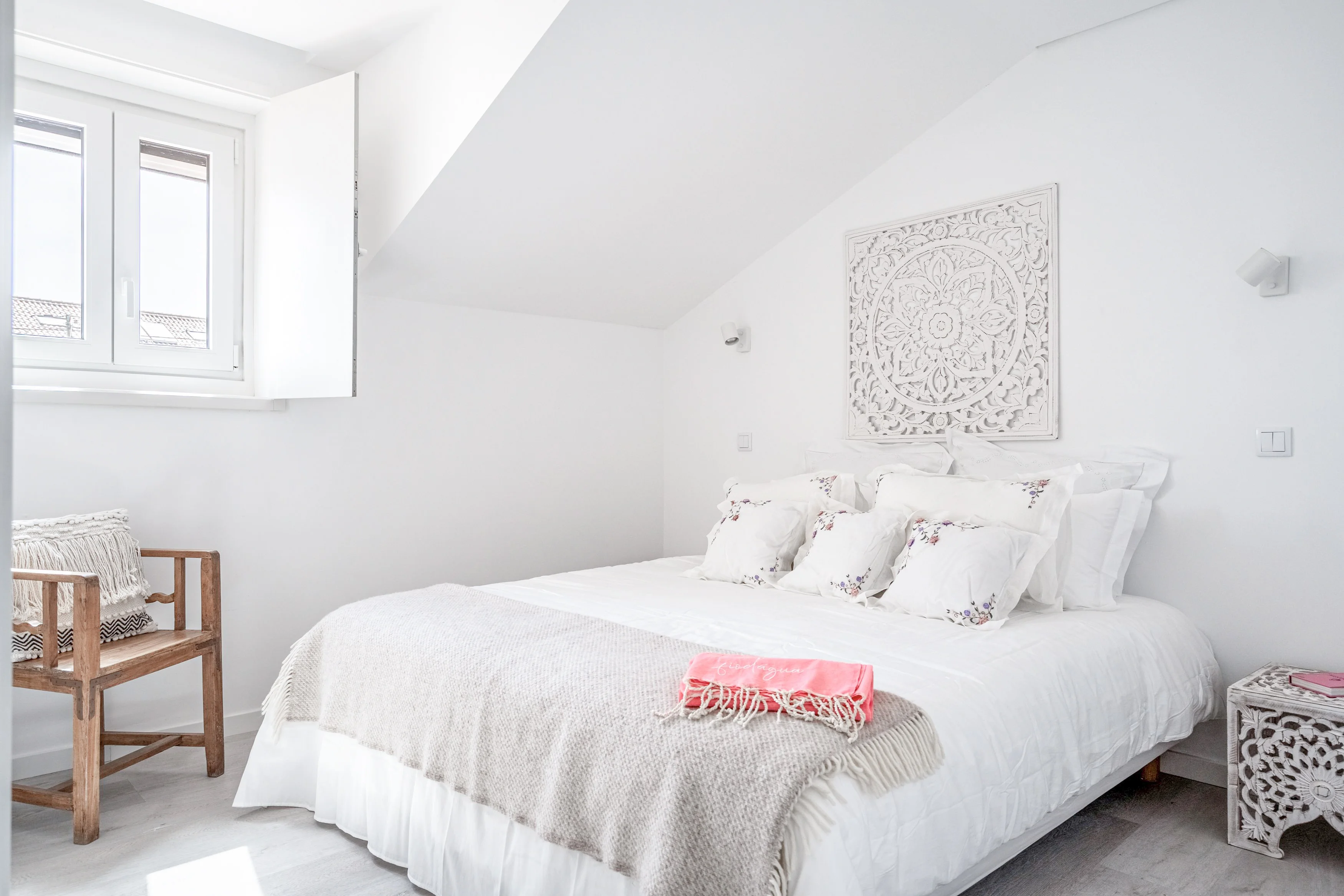 Attic bedroom with sloped ceiling, carved mandala headboard, white linens and view of terracotta rooftops