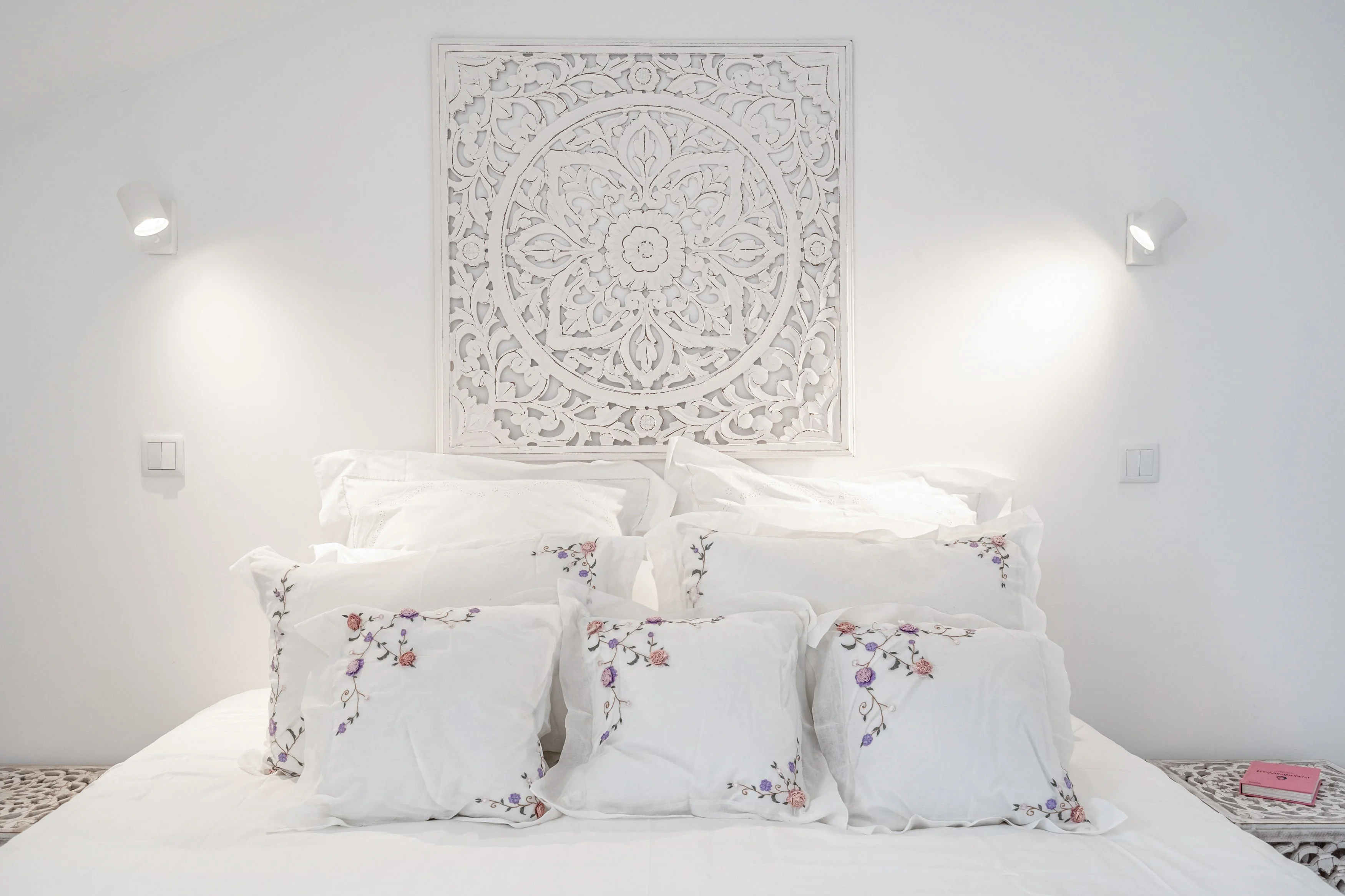 Bed with white linens and decorative pillows with floral embroidery, carved mandala headboard panel and warm wall sconces