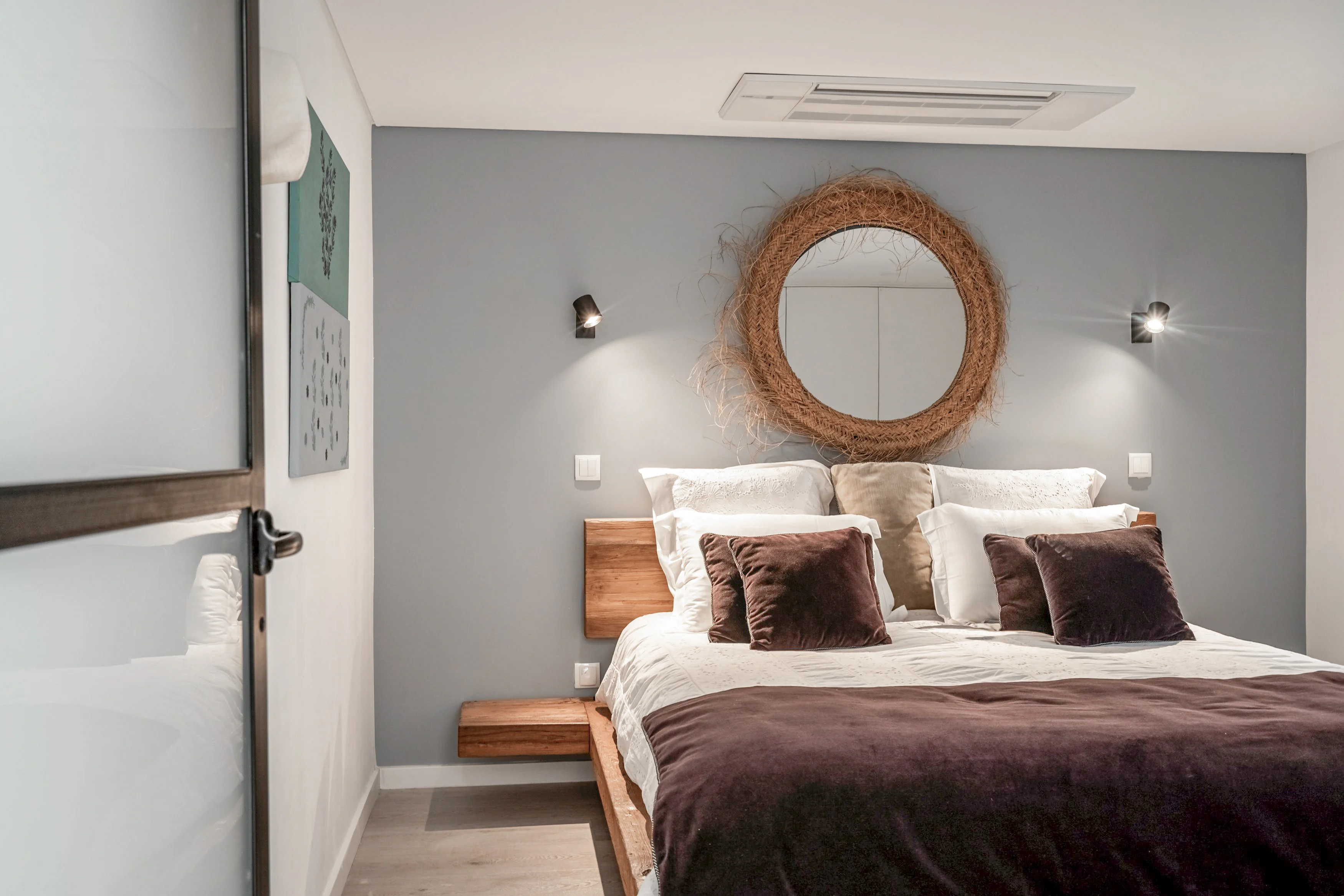 Bedroom with round woven mirror above bed, white linens and reading sconces