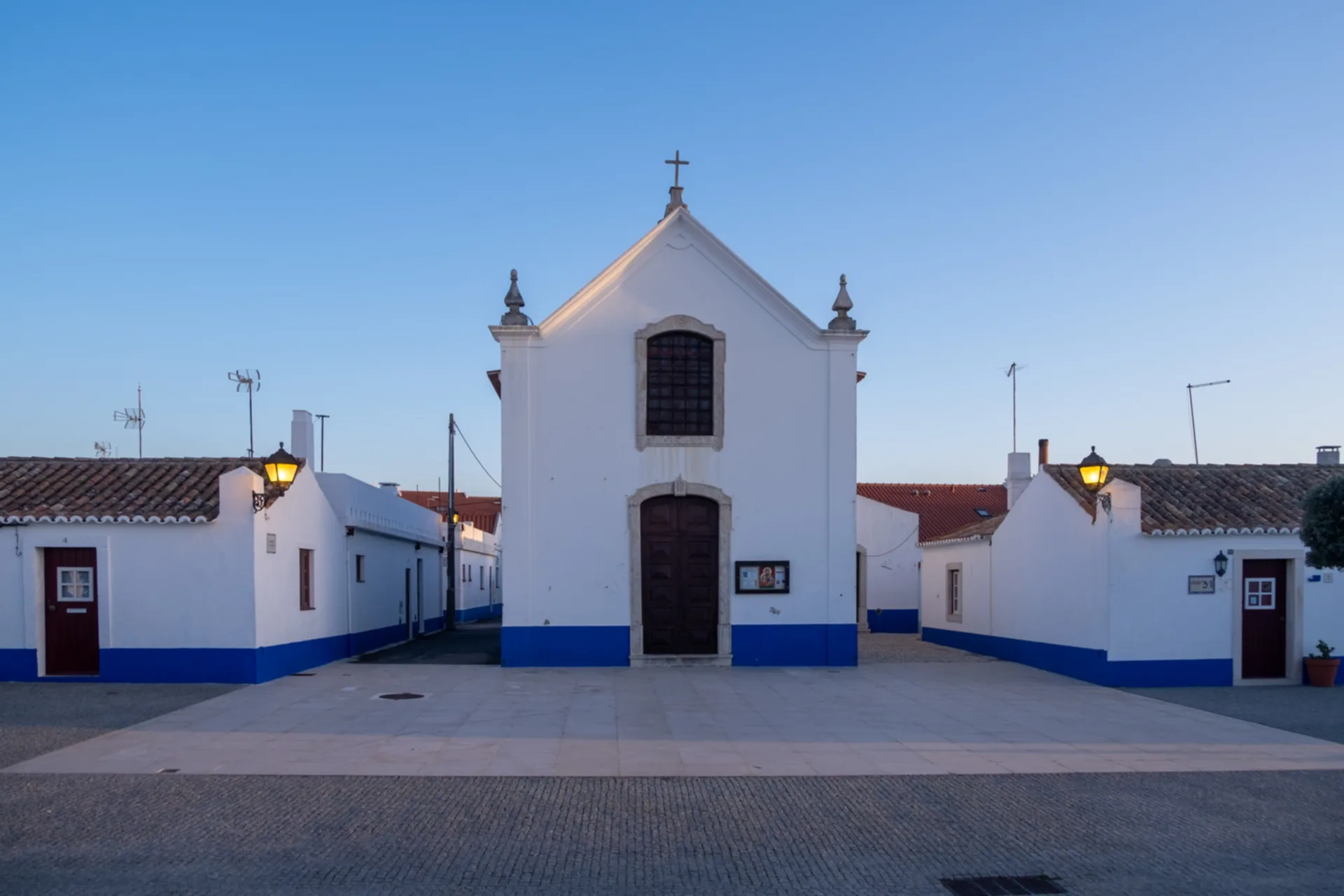 Traditional whitewashed chapel with blue base trim and arched wooden door in a paved stone square, flanked by terracotta-roofed buildings